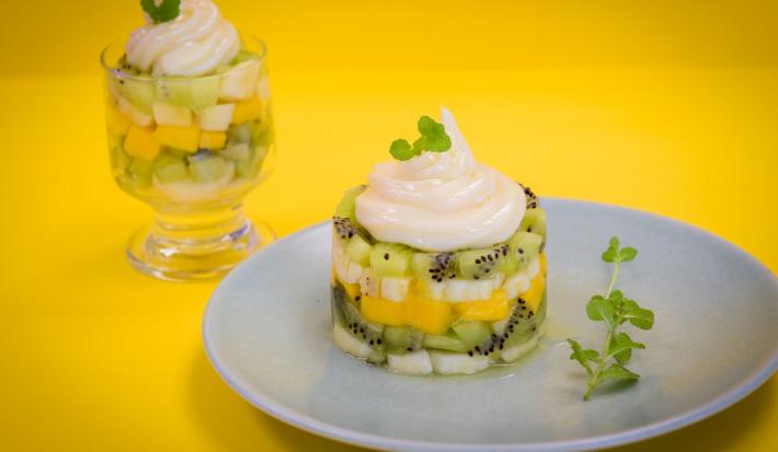 fotografia em tons de amarelo e branco, contém um prato redondo e branco com Tartar com pedaços de frutas e creme por cima e ao fundo uma taça transparente com pedaços de frutas e creme por cima.