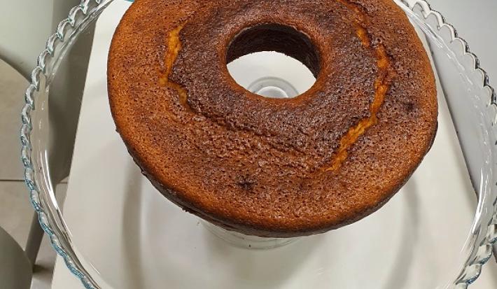 Foto da receita de bolo de cuscuz servida em uma porção grande sobre uma bailarina de vidro em cima de uma mesa branca