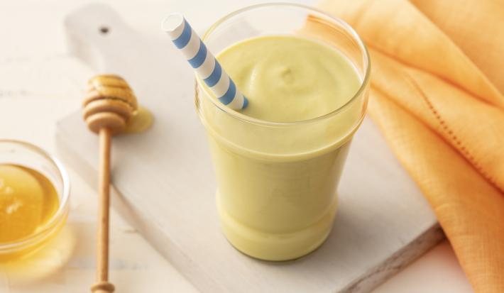 Fotografia em tons de amarelo e verde em uma bancada de madeira clara com um pano laranja, ao centro uma tábua de madeira com um copo de vidro com a vitamina de abacate dentro e um canudo de papel com listras azuis. Ao lado, um potinho de mel.
