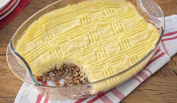 Fotografia em tons de amarelo e branco, ao centro refratário com carne moída e purê de batatas sobre guardanapo branco com listras vermelhas, sobre bancada de madeira.