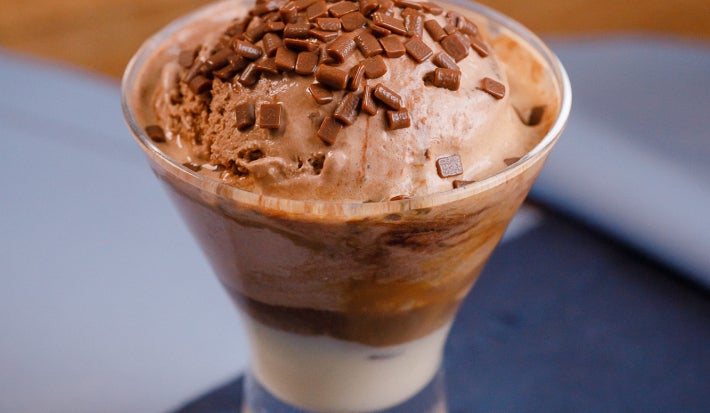 Taça com sorvete de chocolate, raspas por cima e base cremosa, servida sobre tecido azul.