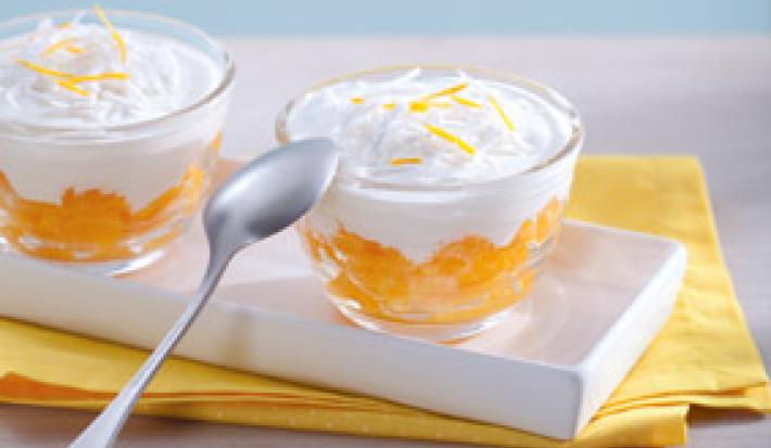 Fotografia em tons de amarelo em uma bancada de madeira clara, um pano amarelo, um recipiente branco retangular com duas taças de vidro com a sobremesa de laranja com creme de coco dentro delas.