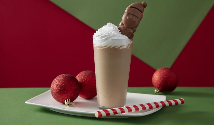 Fotografia em tons de marrom em uma bancada de madeira de cor verde. Ao centro, uma bandeja branca contendo um copo com a bebida dentro. Ao fundo, há 3 bolas de natal.