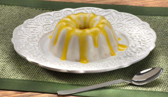 Fotografia em tons de amarelo e verde em uma bancada de madeira com uma toalha verde, um prato redondo branco raso e o flan de coco com calda de pequi em cima dele. Ao lado, uma colher.