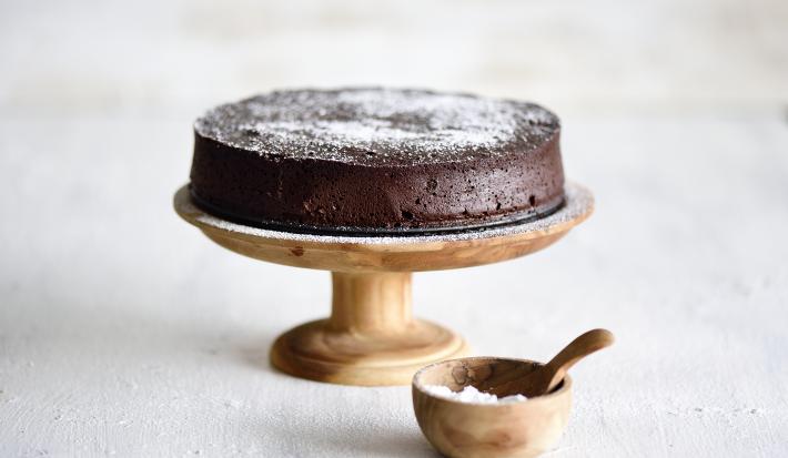 Foto da receita de Bolo Chocolatudo Sem Farinha. Observa-se uma boleira de madeira em um fundo branco com o bolo em cima, polvilhado de açúcar.