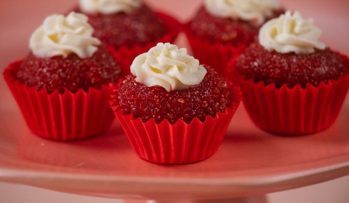 Fotografia de cinco unidades forminhas de cor vermelha, um junto da outra, e dentro de cada forminha tem um brigadeiro de red velvet, passado no açúcar cristal com uma decoração de cream cheese. Os brigadeiros estão sobre um apoio decorativo rosa alto.