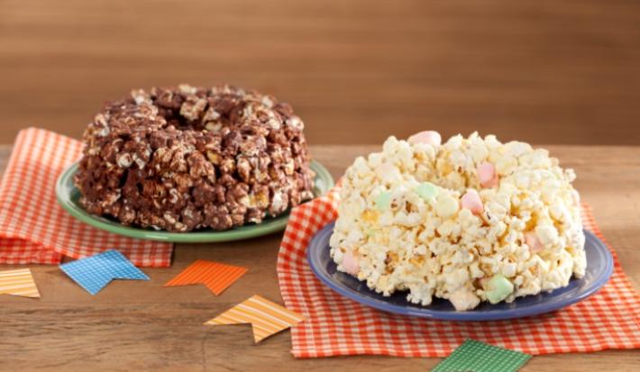 Fotografia em tons de vermelho em uma mesa de madeira com pano xadrez vermelho, um prato verde com o bolo de pipoca de chocolate e outro prato com o bolo de pipoca de chocolate branco.