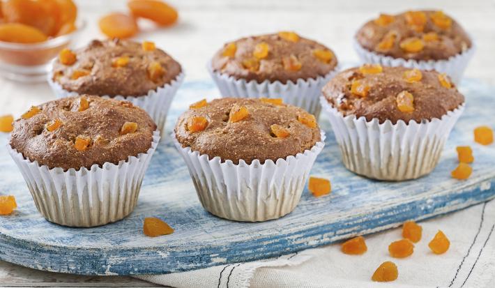 Fotografia em tons de azul em uma bancada de madeira clara, um paninho branco com listras azuis escuras, uma tábua de madeira azul e vários muffins integrais com damasco em forminhas brancas.