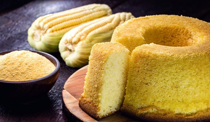 Fotografia de um bolo de milho cortado, revelando seu interior macio, acompanhado de uma tigela de fubá e duas espigas de milho. A cena rústica transmite um apelo caseiro.