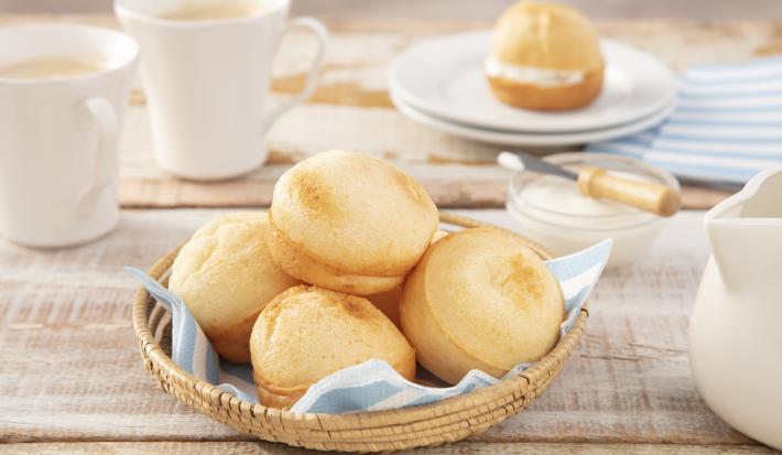 Fotografia em tons de azul em uma bancada de madeira clara, uma cesta de vime com um guardanapo de papel azul e vários pães de queijo dentro dele. Ao lado, um potinho com patê, xícaras brancas e bule de café branco.