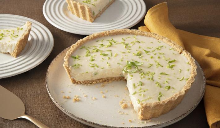 Foto da receita de torta de limão com chandelle servida em uma porção grande sobre uma base de porcelana branca com um pedaço cortado em um pratinho menor ao lado. À frente há uma espátula e ao fundo um pano amarelo