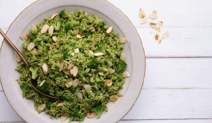 Fotografia em tons de verde em uma bancada de madeira com um prato redondo fundo com o arroz de brócolis e lascas de amêndoas.