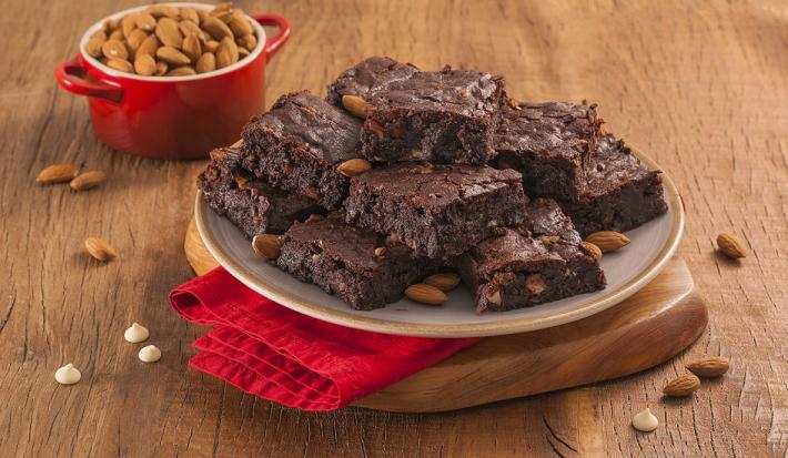 Fotografia em tons de marrom e vermelho de uma bancada de madeira com uma tábua de madeira e um paninho vemelho, sobre ele um prato redondo com pedaços de brownie. Ao fundo um recipiente redondo com amêndoas.