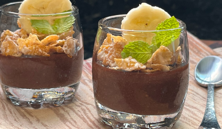 Imagem mostra 2 copinhos transparentes com um creme de chocolate escuro até a metade, seguido por cereais matinais, uma rodela de banana e folhas de hortelã. Os copos estão sobre um pano marrom e uma colher prateada está ao lado direito.