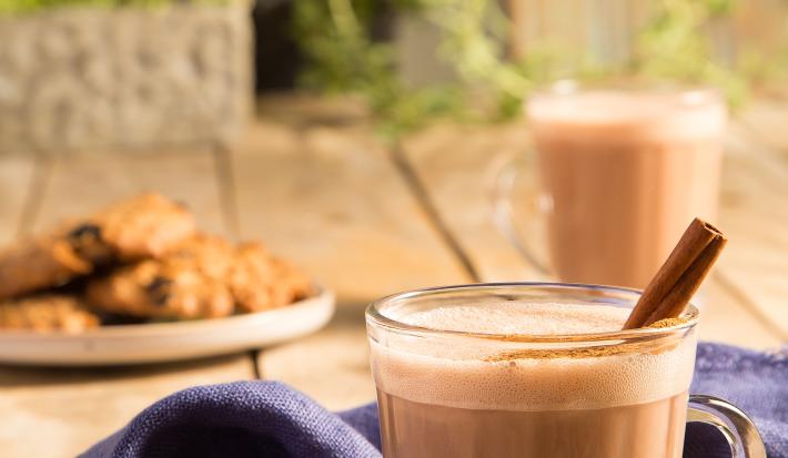Fotografia em tons de marrom em uma bancada de madeira com um pano azul e duas xícaras de vidro com o chocaccino e um pau de canela para enfeitar. Ao fundo, um prato com cookies