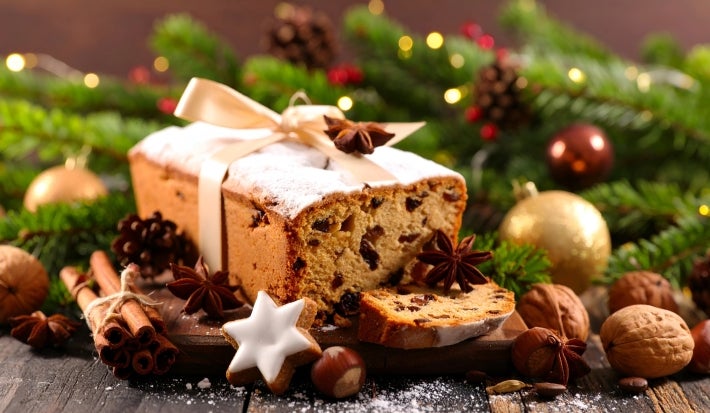 Fotografia de um bolo de frutas decorado com açúcar em pó e um laço, cercado por nozes, anis estrelado e canela, em um ambiente festivo com luzes quentes e ramos de pinheiro.