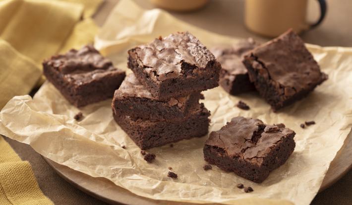 Fotografia em tons de marrom e bege em uma bancada de madeira, um pano bege, uma tábua de madeira redonda, um papel manteiga marrom com vários brownies de chocolate em cima. Ao fundo, xícaras beges.