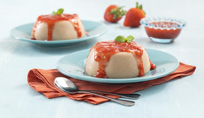 Foto da receita coringa de Flan. Observa-se dois pratos azuis claros com o flan e a calda de morangos por cima, o da frente sobre um guardanapo marrom e ao lado duas colheres de sobremesa. Ao fundo, morangos e a calda em um pote decoram o cenário.