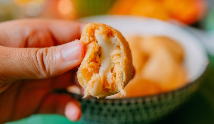 Fotografia de uma mão mostrando metade de uma coxinha aberta de frango e catupiry. Ao fundo tem coxinhas dentro de um recipiente branco e verde fundo.