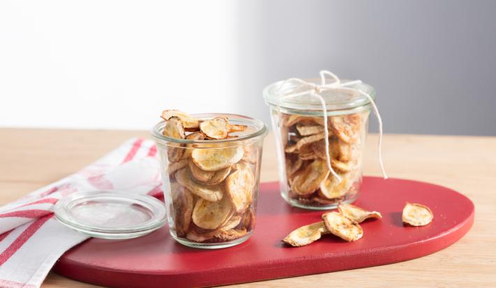 fotografia em tons de bege e vermelho tirada de uma bancada bege. Contém uma tábua vermelha com dois potes transparentes com bananas cortadas em rodelas finas fritas. Ao fundo um pano em tons de branco e vermelho.