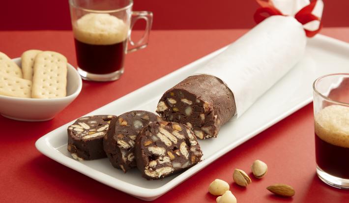 Foto da receita de Choco Salame. Observa-se uma cenografia vermelha com uma tábua branca e o chocosalame por cima, envolvido num papel anteiga. Dois copinhos de espresso decoram a foto.