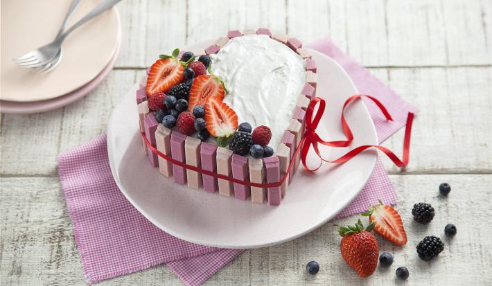 fotografia em tons de branco e rosa de uma bancada branca vista de cima, ao centro um pano rosa com um prato redondo por cima um bolo em formato de coração com marshmallow, morangos e frutas vermelhas por cima para decorar.