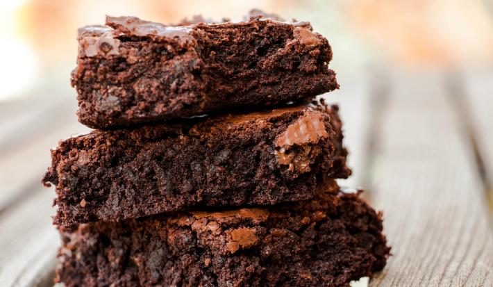 Fotografia de três pedaços de brownie sobrepostos sobre uma mesa de madeira clara.