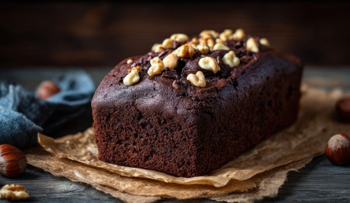 Bolo de chocolate recém-assado, com uma cor rica e escura, coberto com nozes. Ele está sobre papel em uma superfície de madeira rústica.