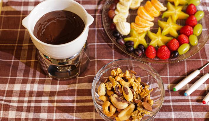 Fotografia em tons de marrom em uma bancada com uma toalha xadrez em marrom, com uma pequena panela branca com o fondue de chocolate dentro, ao fundo um recipiente com frutas picadas e um potinho de vidro com castanhas e amêndoas.