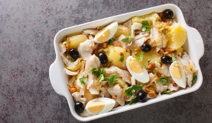 Em cima de uma mesa, refratário com bacalhau, batatas, ovos cortados ao meio e azeitonas pretas, decorado com folhas de manjericão.