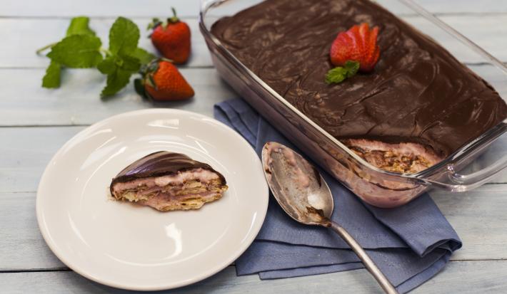 Fotografia em tons de azul e chocolate, com refratário contendo um pavê com chocolate decorado com morango sobre guardanapo azul, no entorno prato com porção do pavê, colher, morangos, folhas de hortelã, e tudo sobre bancada em tons de azul.
