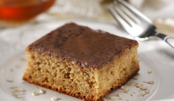 Fotografia em tons de marrom em uma mesa com uma toalha branca e bege, um prato branco raso redondo com uma fatia do bolo de banana com aveia e mel. Ao fundo, um potinho de mel.