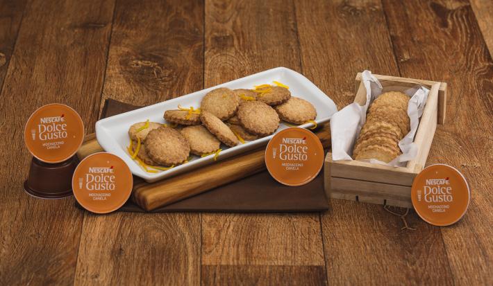 Fotografia em tons de marrom em uma bancada de madeira escura, um pano marrom com uma tábua de madeira em cima dele e um recipiente branco com vários biscoitos amanteigados com raspas de laranja em cima deles. Ao lado, uma caixinha com mais biscoitos.