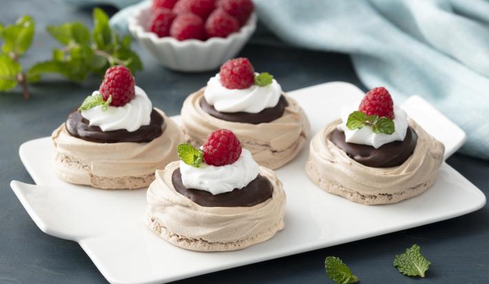 Fotografia em tons de vermelho em uma bancada de madeira azul escura, um pano azul claro, uma tábua branca ao centro com quatro Pavlovas em cima dela, decoradas com framboesa, chocolate e chantilly.