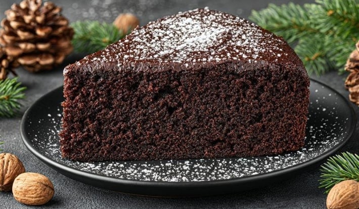 Fotografia de uma fatia de bolo de chocolate escuro em um prato preto, também polvilhada com açúcar de confeiteiro. Elementos como pinhas e nozes ao redor evocam um ambiente rústico e festivo.