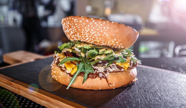 Fotografia de um hamburguer de carne e champignon temperado e servido com rúcula. A fatia do pão na parte de cima, está ao contrário, e o lanhe está sobre uma tábua de madeira com fundo preto.