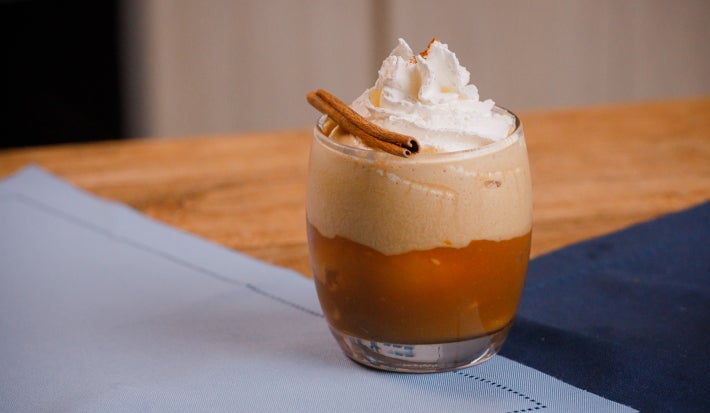 Copo baixo com bebida em camadas, espuma no topo, chantilly e canela em pau sobre tecido claro.