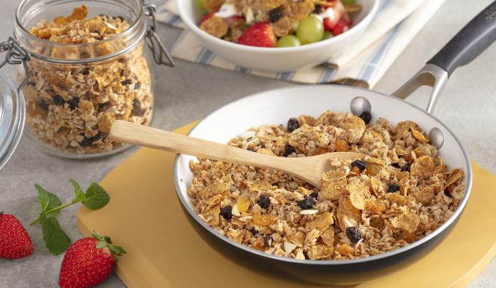 Fotografia em tons de amarelo em uma bancada de madeira com uma tábua de madeira, uma frigideira grande dentro com a granola com uvas-passas, cereal matinal dentro. Ao lado, um potinho com a granola e ao lado um outro potinho branco com frutas.