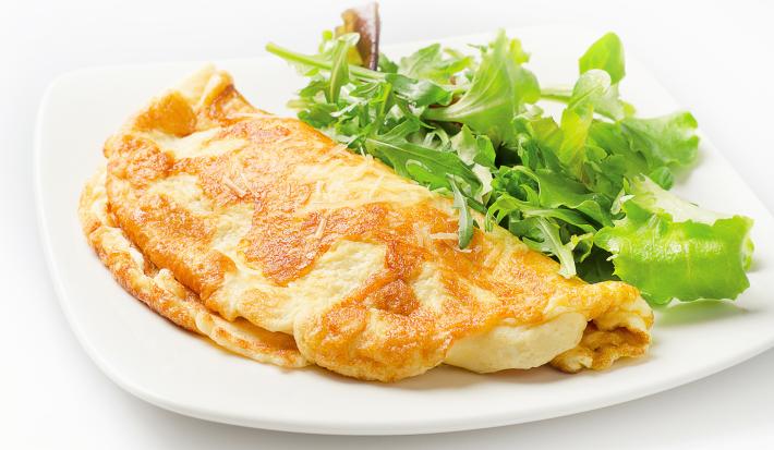 Fotografia de um omelete com crosta de queijo crocante dobrado ao meio, ao lado de uma salada de folhas, sobre um prato de vidro branco. O prato está sobre uma mesa branca.