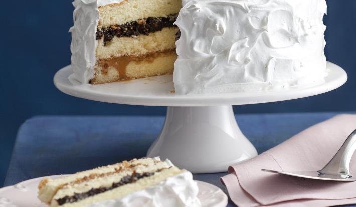Fotografia em tons de branco e azul, ao fundo, sobre bancada azul, uma boleira de porcelana com bolo fatiado coberto com marshmallow e recheio de ameixa, prato de sobremesa com bolo de ameixa, dois pratos com talheres e um guardanapo com uma espátula.