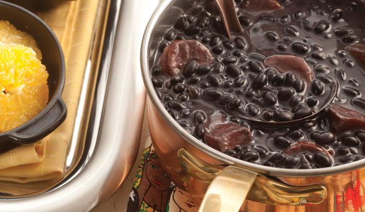 Fotografia em tons de bege em uma bancada de madeira clara com um suporte para panela colorido com rostos de mulheres, uma panela dourada em cima dele com a feijoada dentro.