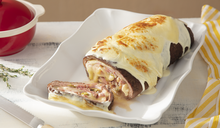 Foto em tons de branco da receita de rocambole de carne moída recheado servida em uma base de porcelana branca com um pedaço cortado. Ao lado há um pano amarelo listrado e uma panela vermelha, além de uma faca de cortar carne