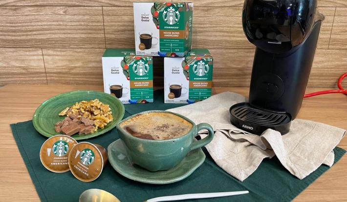 Fotografia em tons de verde e bege em uma bancada de madeira, um paninho bege, uma caneca verde com o petit gateau com café dentro. Ao fundo, máquina dolce gusto, e cápsulas de dolce gusto house blend.