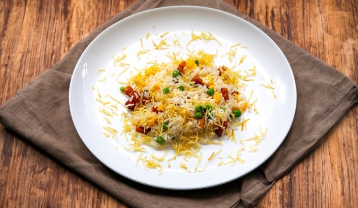 Em um prato branco redondo, porção de arroz com ervilha, pedaços de bacon e batata palha.