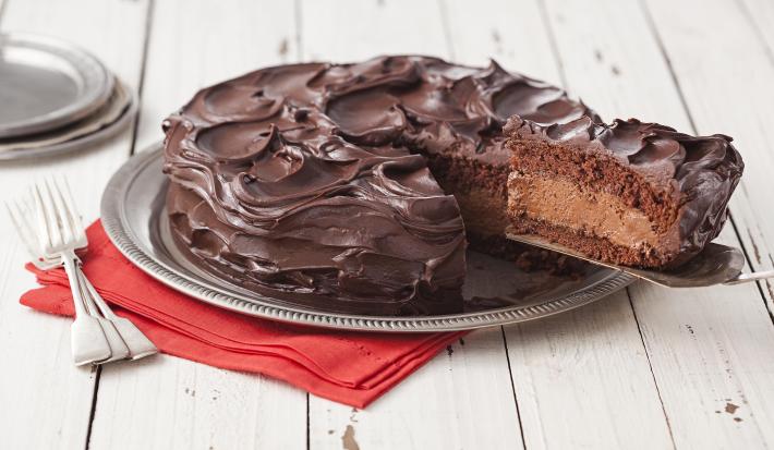Fotografia em tons de branco e marrom de uma bancada de madeira, um paninho vermelho, um prato redondo prateado, dois garfos prateados e um bolo de chocolate com uma fatia sendo retirada, mostrando o recheio de mousse.