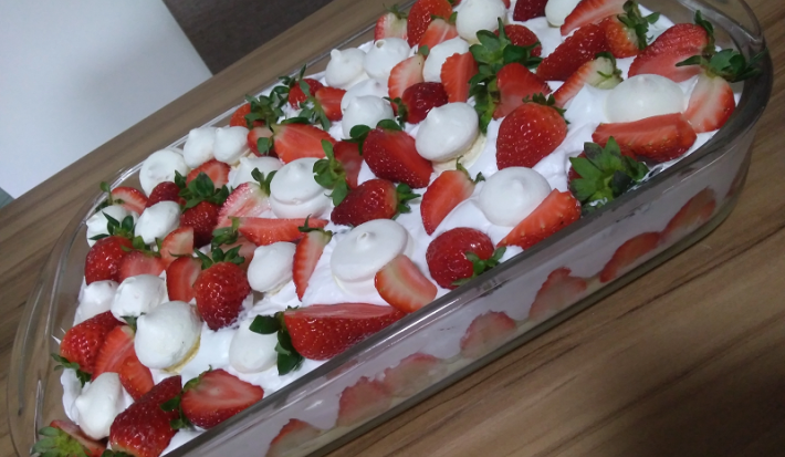 Foto em tons de vermelho da receita de merengue de morango com suspiros servida em uma travessa de vidro grande com morangos e suspiros por cima decorando. A receita está sobre uma mesa de madeira