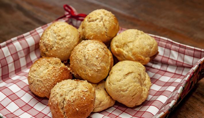 Fotografia de pãezinhos com massa de requeijão e queijo parmesão. Os pães estão dentro de uma cestinha quadrada com papel quadriculado vermelho e branco, que está sobre uma mesa de madeira.