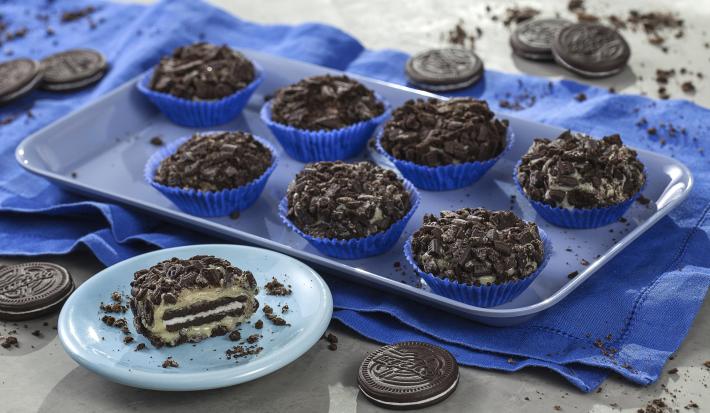 fotografia em tons de azul, preto e cinza de uma bancada cinza vista de cima, contém uma bandeja azul com forminhas azuis e brigadeiros de biscoitos e abaixo da bandeja um pano azul. Um pratinho na frente azul e redondo com uma fatia de brigadeiro.