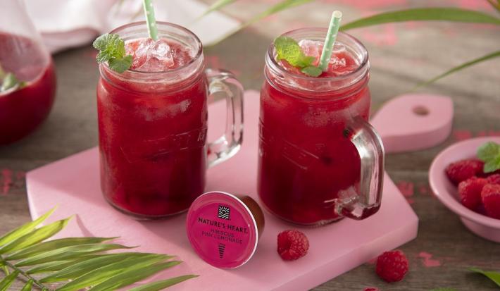 Foto em tons de cor de rosa da receita de pink lemonade com frutas vermelhas e hortelã servida em canecas altas de vidro em cima de uma tábua rosa clarinha com um pote de framboesas ao lado