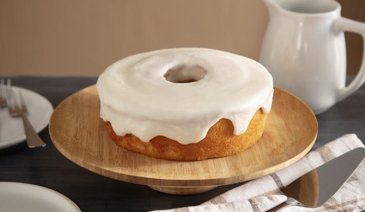 Foto em tons claros da receita de cobertura de porcelana de coco servida sobre um bolo em cima de uma base redonda de madeira. Ao lado há um prato de porcelana branco com talheres prateados e um pano bege à frente
